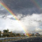 Carretera en día lluvioso con nubes muy oscuras, gran contraste lumínico y un arcoíris en el horizonte (hacia la izquierda).