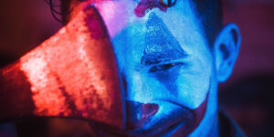 Imagen de un hombre con maquillaje de payaso, mirada torva e iluminación azul, que cubre media cara con la hoja de un hacha iluminada en rojo.
