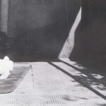 Fotografía en blanco y negro de un bebé de apenas un par de meses, vestido de blanco, sin pelo en la cabeza y en el suelo de un patio, sobre una alfombrilla.