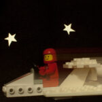 Un astronauta de LEGO de color rojo viaja en una pequeña nave sobre un fondo de estrellas que podría haber recortado un niño.