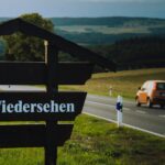 Un coche se aleja por una carretera en la campiña, en segundo plano. En primer plano, un cartel con la inscripción "Auf Wiedersehen".