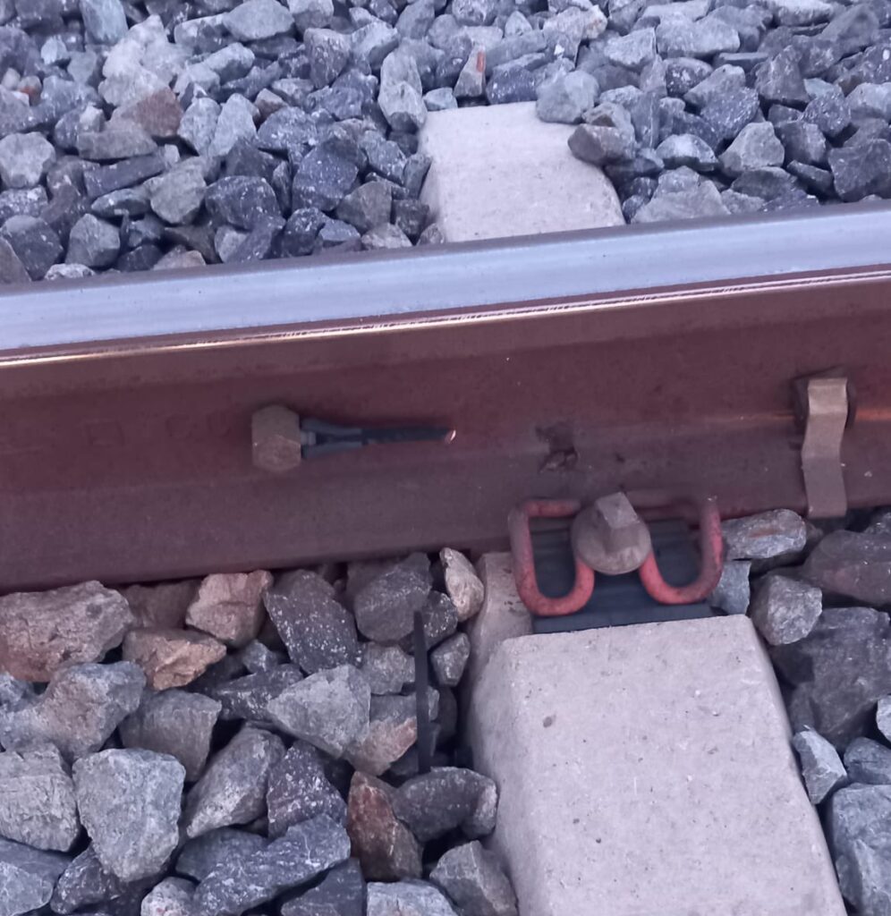 Un raíl ferroviario sobre una traviesa, por el lado interior. Se aprecia la conexión con el cable del circuito de vía, cortada.