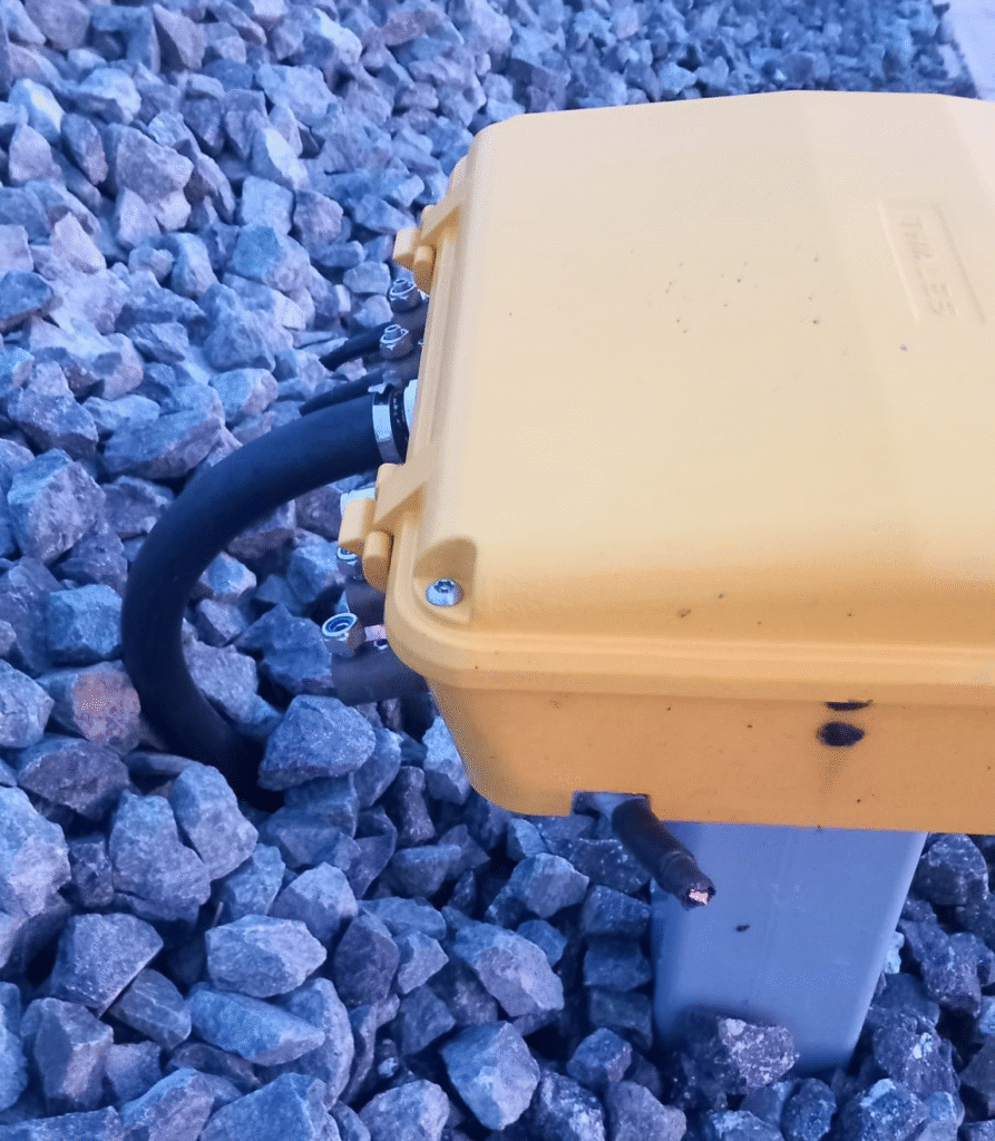 Caja de conexiones de circuitos de vía, sobre una peana metálica anclada en el balasto, una caja amarilla de la que salen varias mangueras de cable. Algunas están cortadas.