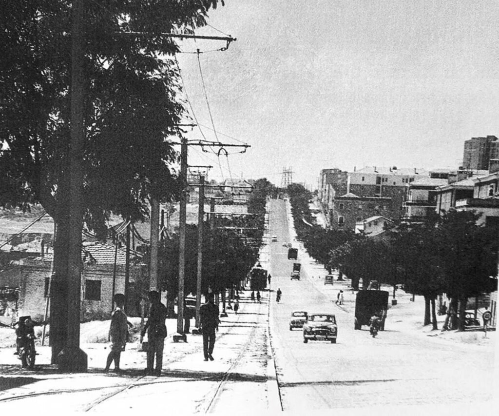 Vista de la carretera de Extremadura en 1964. El centro de Madrid queda a la espalda. Se ve una vía de tranvía, única, con un tranvía al fondo. A su derecha, una calle asfaltada relativamente ancha para tener solo un carril por sentido, probablemente adoquinado. En los márgenes hay árboles y, más lejos, casas bajas (a la izquierda) y edificios de tres o cuatro alturas (a la derecha). Apenas se aprecian un total de diez vehículos, aunque la vista permite apreciar alrededor de un kilómetro hacia el horizonte.