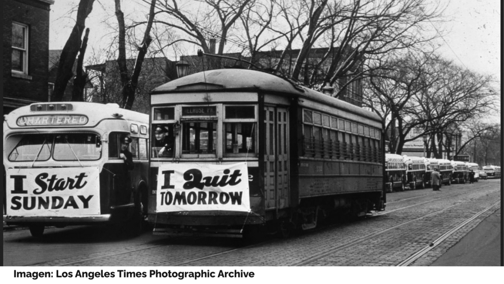 Un autobús y un tranvía juntos en una calle. El autobús lleva una pancarta con el mensaje "I start Sunday", y el tranvía otra muy similar, que reza "I quit tomorrow". Imagen: Los Angeles Times Photographic Archive.