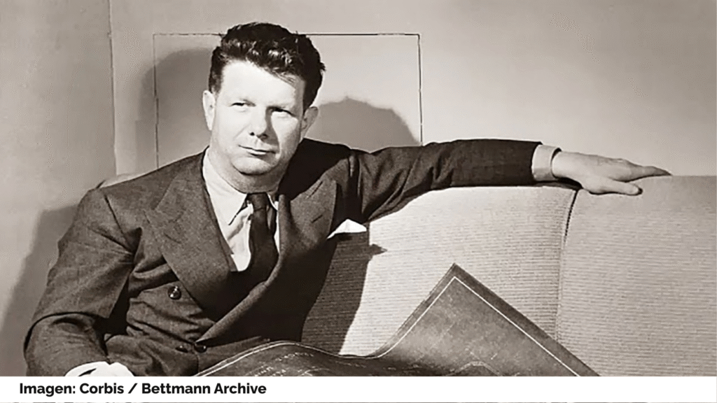 Retrato de Norman Bel Geddes. Lleva americana cruzada y corbata. Consulta unos planos sentado en un sofá. Imagen: Corbis, Bettmann Archive.