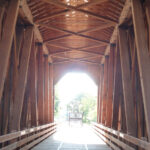 Interior de un puente cubierto de madera, antaño de uso ferroviario y actualmente peatonal. Se aprecia la estructura de vigas cruzadas en paredes y techo. La salida, en el centro de la imagen, es un dintel con las esquinas achaflanadas. Entra mucha luz.