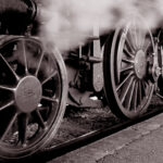 Locomotora de vapor checa fotografiada desde abajo y atrás, por el costado derecho, hacia adelante con un ángulo bastante cerrado. Se aprecian seis ruedas, de las que las cuatro centrales son tractoras, unidas por bielas. Fotografía en blanco y negro.