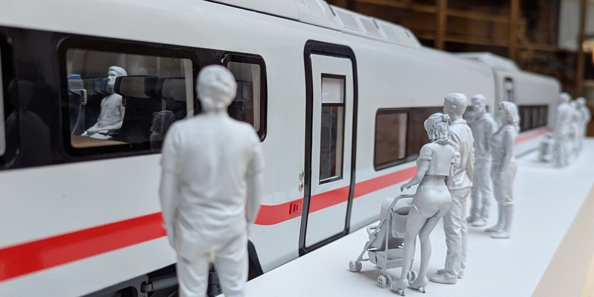 Maqueta de un ICE-L de Talgo con librea blanca con banda roja de la DB. Varias figuritas humanas pintadas totalmente de blanco esperan en un andén a que se abran las puertas para acceder.