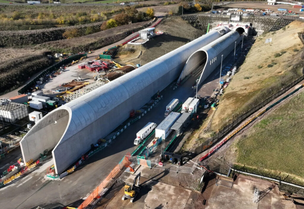 Trabajos de construcción en la salida de un túnel ferroviario de doble tubo vistos desde el aire. Se aprecian dos falsos túneles frente a las bocas reales que hacen de visera, con los extremos biselados hacia arriba y con perforaciones a intervalos regulares en los laterales.